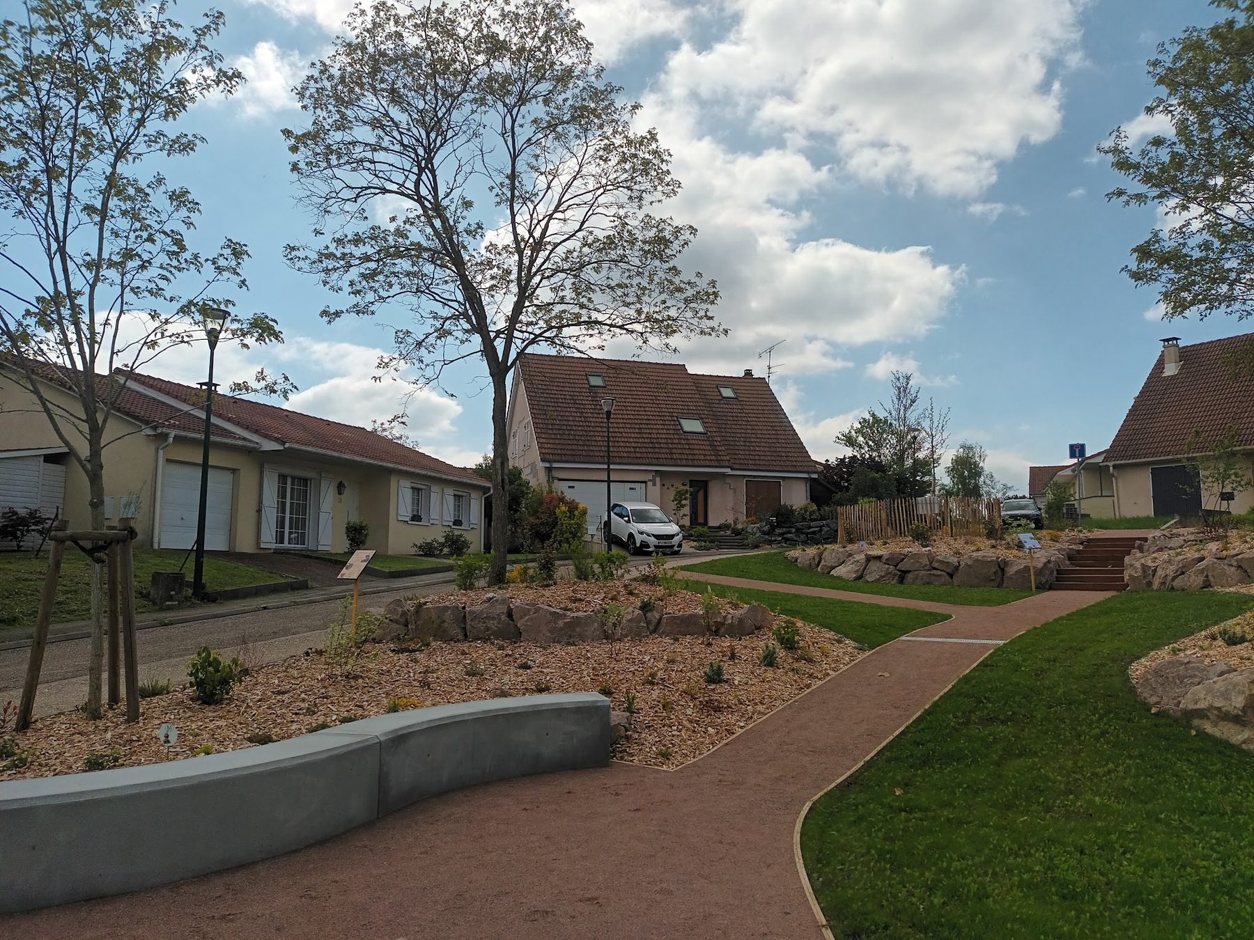Espace vert aménagé avec allée, enrochements, banc en béton et plantations